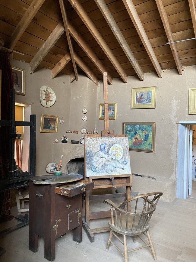 Interior view of the Eanger Irving Couse Studio, Taos, NM Interior view of a chair seated in front of an easel with a painting. Atleft is a small table with an open drawer and an easel on its surface. In the background is a corner fireplace around which hang framed paintings.