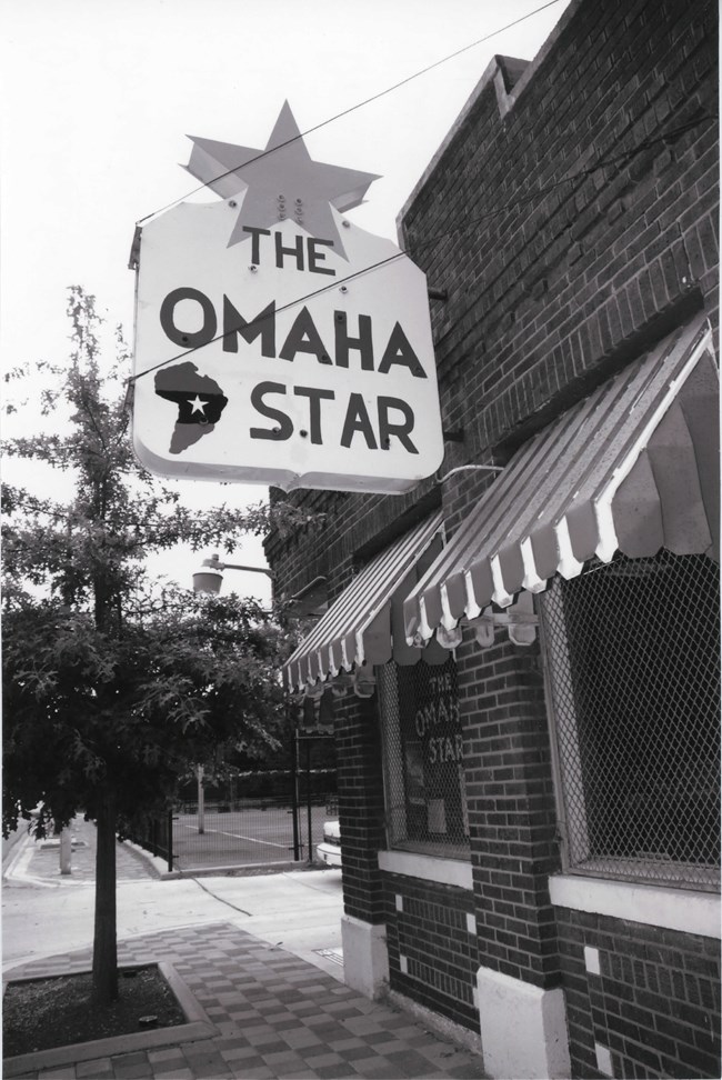 Small brick building on street corner with large sign stating: The Omaha Star with an image of Africa in the sign