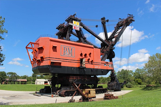 100001945 Big Brutus (KS) view Image of gigantic mining excavator, set among gravel, grass and trees, with smaller equipment in the foreground and buildings in the background.