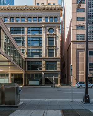 A six story historic building made of light-brown brick sits along a street in a downtown area.