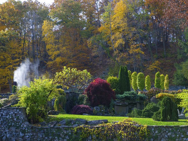 Park setting with colorful trees, stone wall, and water spout in the background