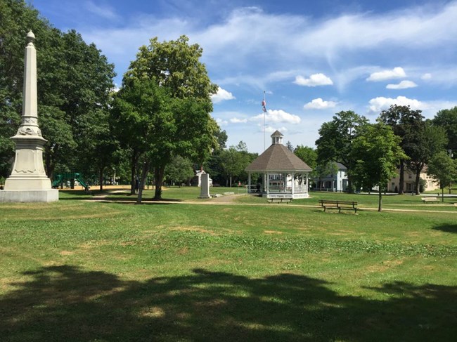 Green park square with a white gazebo, monolith, and park bench, with trees and walking paths