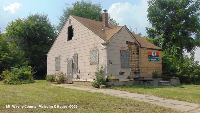 100007205_MalcolmX_image An eroded paved path leads through the grass to a tan house with wood boards covering the damage from a side view.