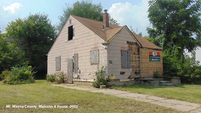 An eroded paved path leads through the grass to a tan house with wood boards covering the damage from a side view.