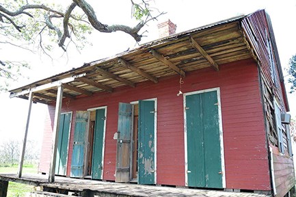 A weathered red wooden building with a porch and peeling green doors, under a large tree branch.