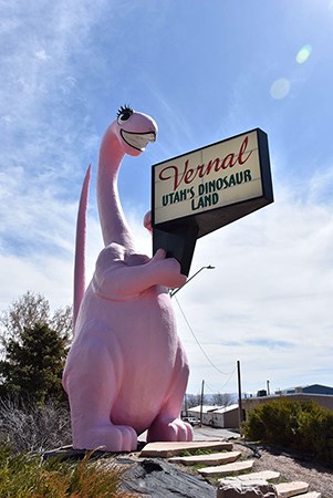 100009526 Dine-a-Ville dinosaur view Image of a pink anthropomorphic dinosaur holding a sign, rocks, and plants, with buildings in the background.