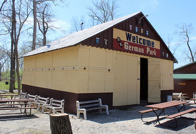 A rustic wooden pavilion with a peaked roof and an open door, featuring a large sign that reads “Welcome German Park.” Picnic tables and benches are arranged outside under leafless trees on a sunny day.