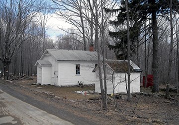 Image of white one-story building, schoolyard, stone wall, vegetation, and road.