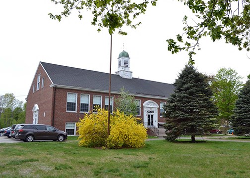 Image of brick building, trees, lawn, and cars.