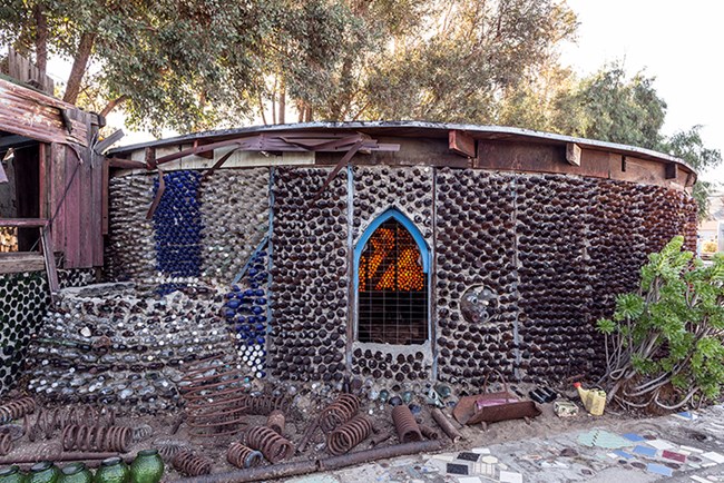 Bottle Village LOC Highsmith Image of a round single story building constructed of colorful bottle, large metal springs, mosaic walkway, and plants.