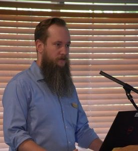White man with a long brown beard wearing a blue button-up shirt stands at a microphone.