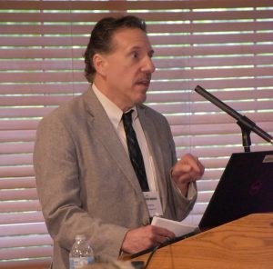 White man with short dark hair wearing a tan jacket, cream shirt, and black tie standing at a podium.