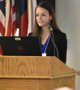 A woman with long brown hair wearing a black jacket and blue shirt stands behind a podium holding a laptop computer.
