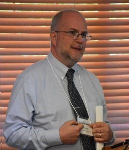 White male with very short gray hair, beard, mustache, and glasses wearing a light gray shirt, polka-dotted black tie, and name tag.