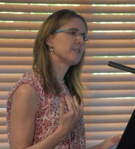Claudia Chemello White woman with long sandy brown hair and glasses wearing a sleeveless res and white leaf pattern top. Standing at a microphone.