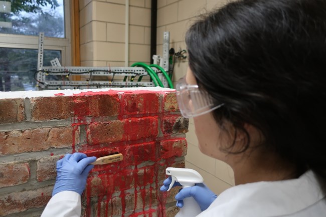 Cleaning Fire Chemical From Brick A person with a bristle brush in their left hand and a spray bottle in their right facing a brick wall with red dye on it.