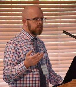 Daniel Phoenix Bald white man in glasses wearing a bright colored plaid shirt and blue necktie standing at a podium.