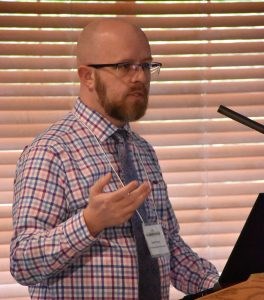 Bald white man in glasses wearing a bright colored plaid shirt and blue necktie standing at a podium.