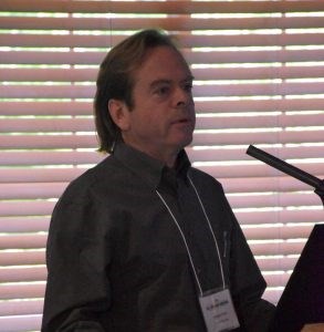 White male with collar length brown hair wearing a dark gray shirt standing at a microphone.