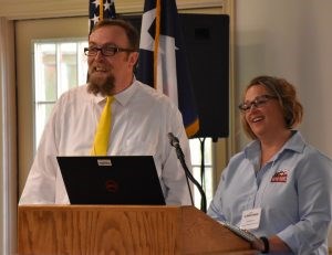 White man in a white shirt and yellow tie and a white woman in a pale blue shirt standing behind a podium.