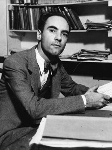 White male in gray suit and bow tie sitting at a desk with a bookshelf behind him.