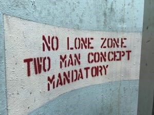 No one allowed past this point alone Pale blue wall with red stenciled text reading no lone zone two man concept mandatory.