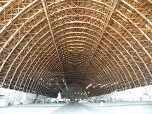 Looking down a long aide open hangar with a semicircular wooden roof supports forming an intricate geometric design.