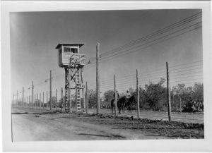 Two men on horseback outside a tall wire fence near a wooden guard tower.