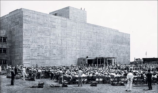 A large crowd sits in front of a multi-story wide rectangular building with a geometric façade.