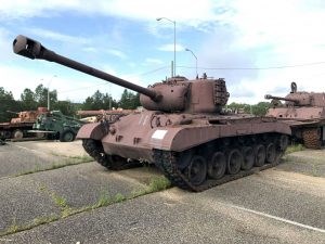 A faded maroon M26 Pershing tank sits on a cement tarmac with other tanks behind it.