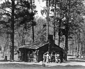 Historic photo – five people in front of a log and stone cabin surrounded by tall pine trees.