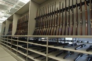 Over 80 historic rifles standing vertically in racks above four long shelves of evenly spaced other barreled weapons.