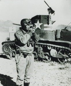 Black and white photo of man coveralls wearing a leather helmet, goggles, and binoculars standing near a M3 tank.