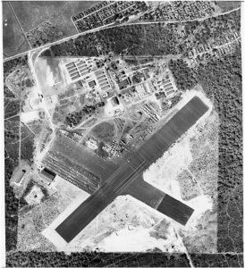 Black and white aerial view looking straight down on X shaped runway in large white square and nearby buildings.