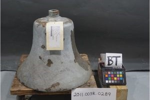 A large weathered bell with tag sits on a wooden pallet with a sign reading 2001.003R.00289. A second sign reads B T.