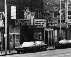 Clubs and bars line a city street in a 1970’s era black and white photo.