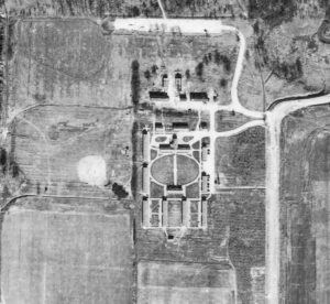 Black and white aerial view of buildings and roads form a rectangle in the center of a field with a larger road on the right.