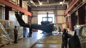 Four people standing inside a large building as the rotor-less UH-1B Huey helicopter is being moved.