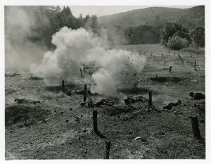 Soldiers crawl under wires as smoke rises from small explosions. Trees and mountains in the background.