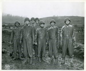 Seven soldiers wearing helmets covered in mud from the neck down standing in a muddy field.