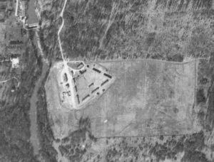 Black and white aerial view of 11 buildings forming a rough triangle in an open field surrounded by a forest.