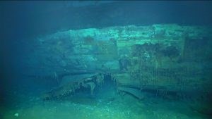 Color image in shades of blue and green of metal hull with hole it in resting on ocean floor.