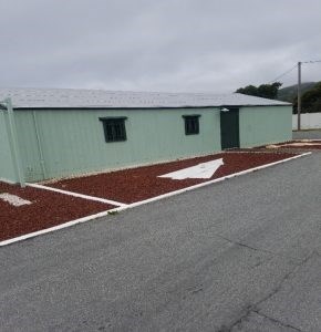 Brown mulch fill large rectangular beds surround the base of a pale green long rectangular building with a gray roof.