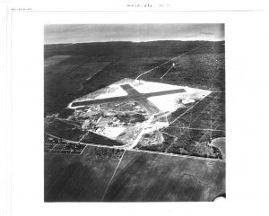 Black and white distance aerial view of X shaped runway in large white square surrounded by gray land.