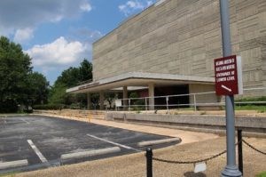 Parking lot and entrance to a multi-story wide rectangular building with a geometric façade.