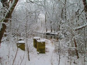 Four short cement rectangular columns in over-grown brush covered in a blanket of snow with a creek in the background.