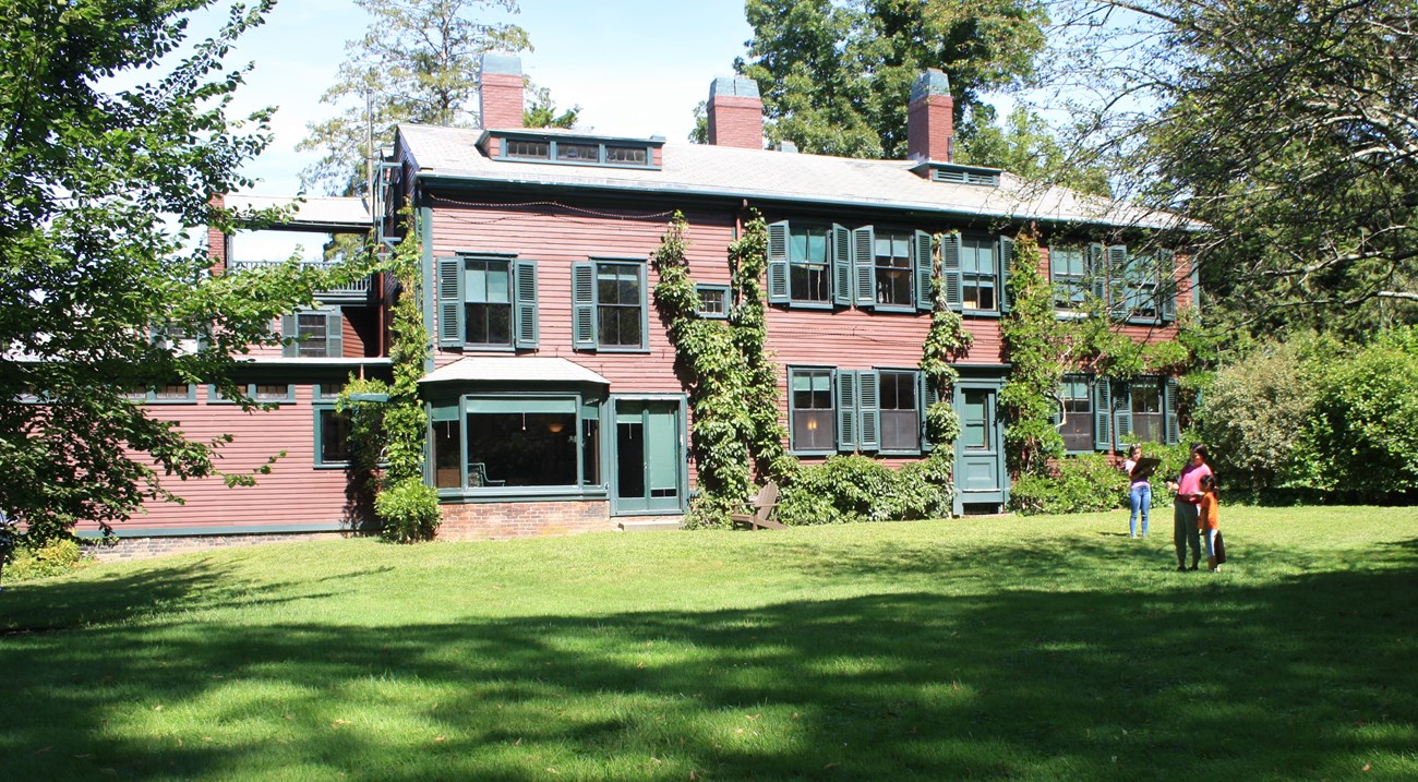 Brown two story home with green trim is surrounded by trees and lawn.