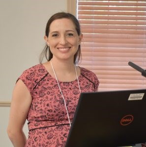 A smiling white woman wearing short sleeve pink top with a black flock design standing behind an open laptop.