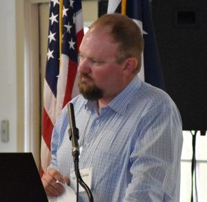 White male with short brown hair mustache and goatee wearing a light blue button-up shirt standing by a microphone.