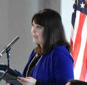 White woman with long dark hair wearing a bright blue jacket over a black shirt standing in front of a microphone.